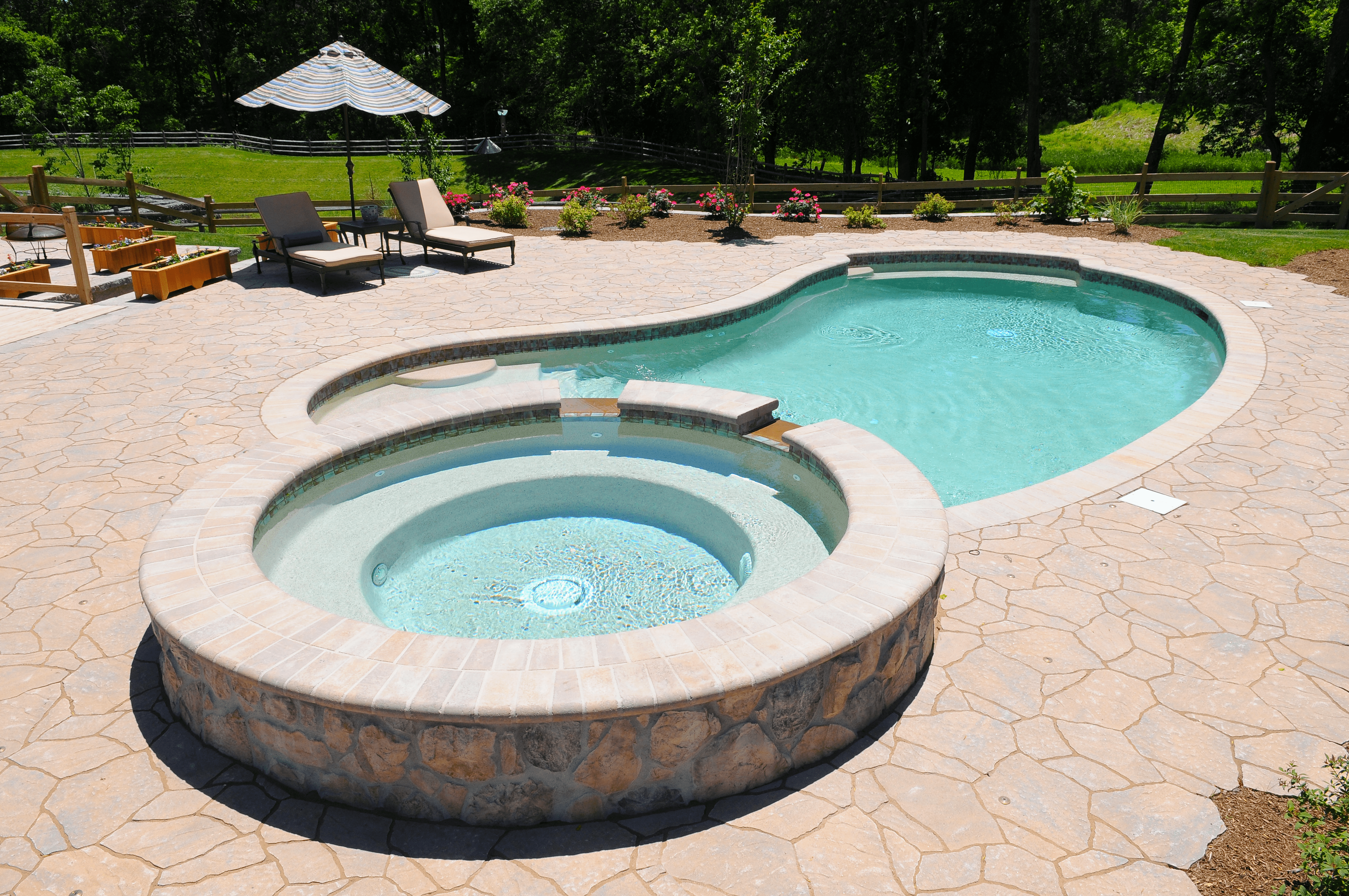 A free-form swimming pool with a light blue interior and a connected circular spa. The spa and pool are surrounded by a textured flagstone patio. A small waterfall feature connects the spa to the main pool. Two lounge chairs with a striped umbrella are on the patio, and a garden with raised wooden planters is visible in the background, along with a wooden fence and trees.