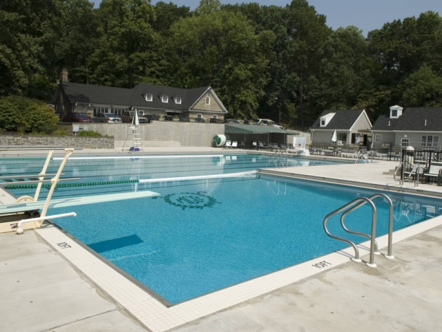 A large, rectangular outdoor swimming pool with lap lanes and a diving board at the far end. The pool is surrounded by a clean, light-colored concrete deck. A handrail provides access to the pool on the right.