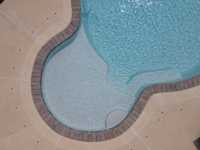 An aerial, close-up shot of the shallow end of a swimming pool. The pool has a light blue interior with a built-in bench and a set of stairs. The edge of the pool is lined with a dark, brick-like coping. The surrounding deck is a light brown, stamped concrete.