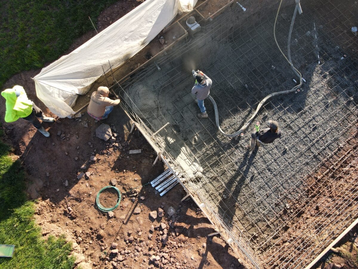 An aerial view of an in-ground swimming pool under construction. The rectangular pool has a rebar framework and is currently being sprayed with shotcrete by a worker inside. Several other workers are visible around the perimeter, and a light-colored tarp is covering a section of the ground. The pool is surrounded by wooden forms and a muddy construction site.