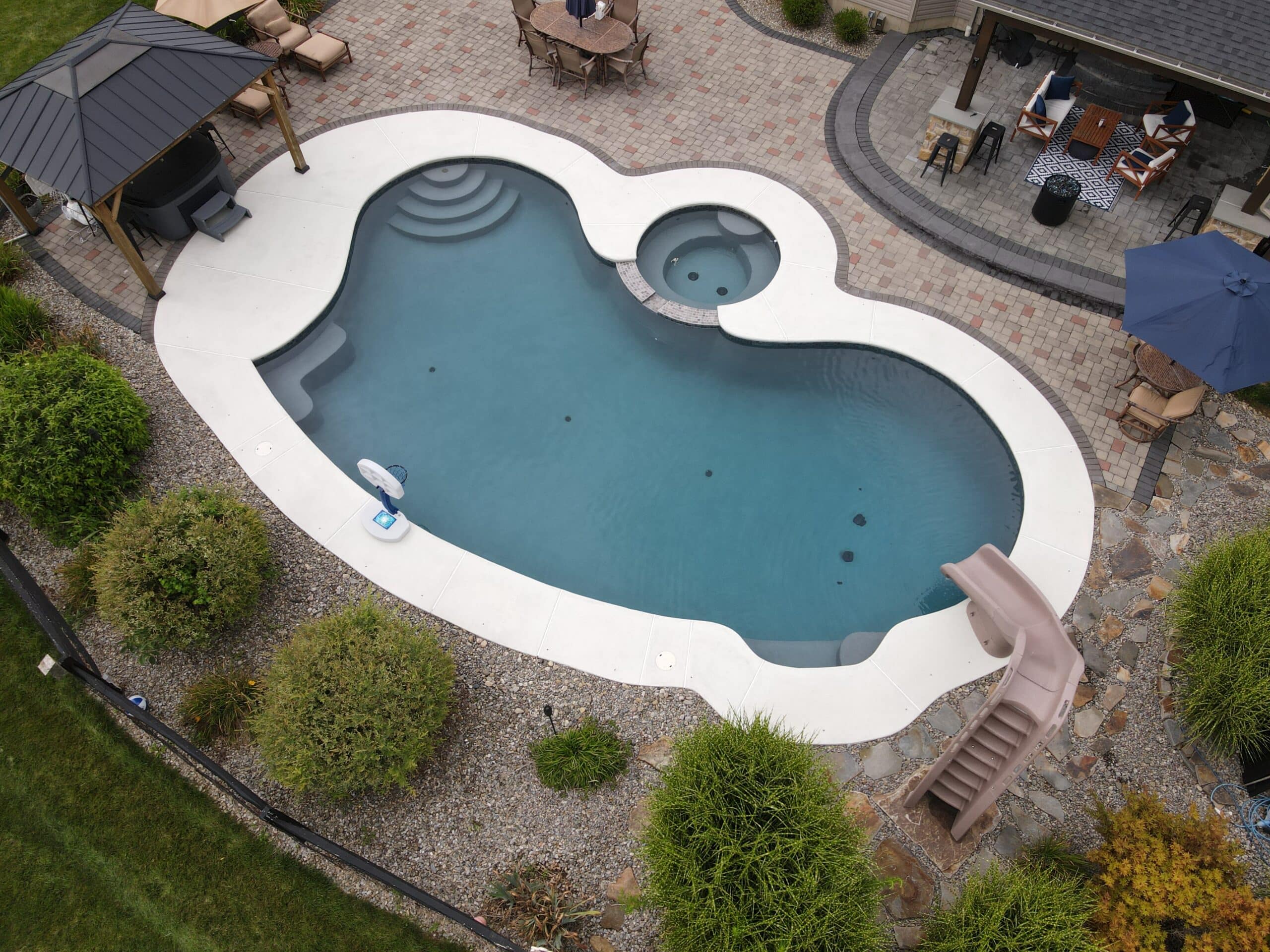 An overhead view of a large, custom-shaped backyard swimming pool with a connecting hot tub. The surrounding patio is a light concrete. The pool area features a hot tub, a built-in slide, a basketball hoop, and several seating areas, including a covered gazebo and a patio dining set.