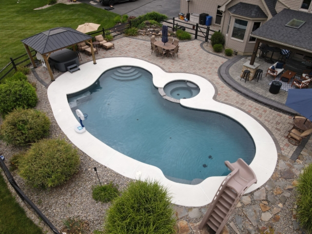 An overhead view of a large, custom-shaped backyard swimming pool with a connecting hot tub. The surrounding patio is a light concrete. The pool area features a hot tub, a built-in slide, a basketball hoop, and several seating areas.