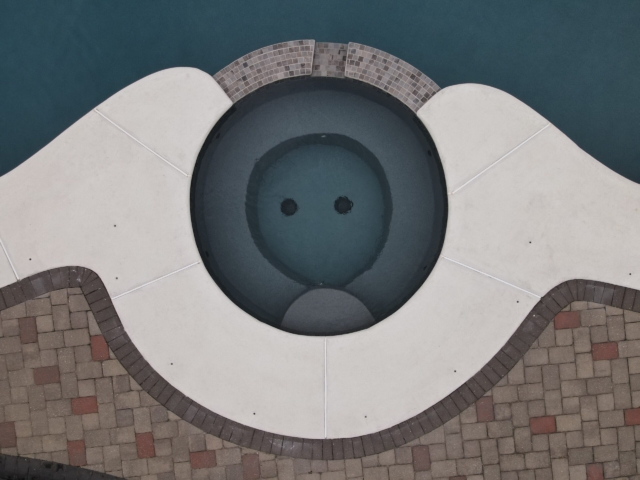 An overhead, close-up shot of a circular hot tub connected to a swimming pool. The hot tub is surrounded by a white concrete deck with a stone tile inlay. The surrounding deck is made of multi-toned pavers in a herringbone pattern.