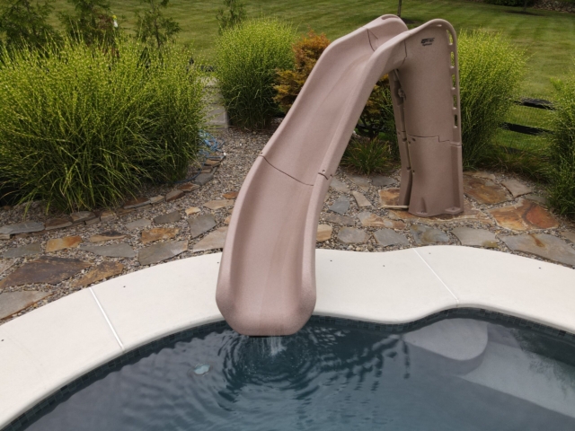 A beige-colored, curved water slide empties into a swimming pool with a dark tile border. The pool's coping is a smooth, light-colored material. The area behind the slide is landscaped with multi-toned flagstone pavers, gravel, and various ornamental grasses.