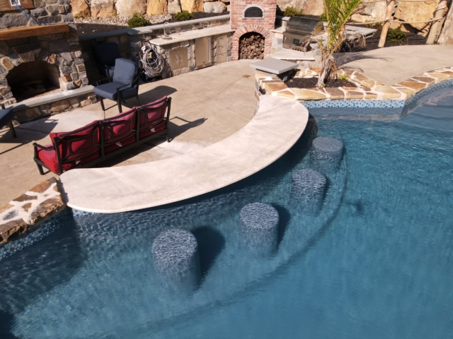 An side shot looking at an outdoor swimming pool with a swim-up bar. The pool has a light blue interior and is surrounded by a light-colored patio deck. A curved, white bar top separates the pool from the deck, and four gray circular bar stools are submerged in the water. The pool is bordered by a wall of natural stones on one side, and a few red lounge chairs are on the deck.