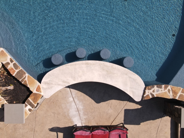 An aerial shot looking down at an outdoor swimming pool with a swim-up bar. The pool has a light blue interior and is surrounded by a light-colored patio deck. A curved, white bar top separates the pool from the deck, and four gray circular bar stools are submerged in the water. The pool is bordered by a wall of natural stones on one side, and a few red lounge chairs are on the deck.