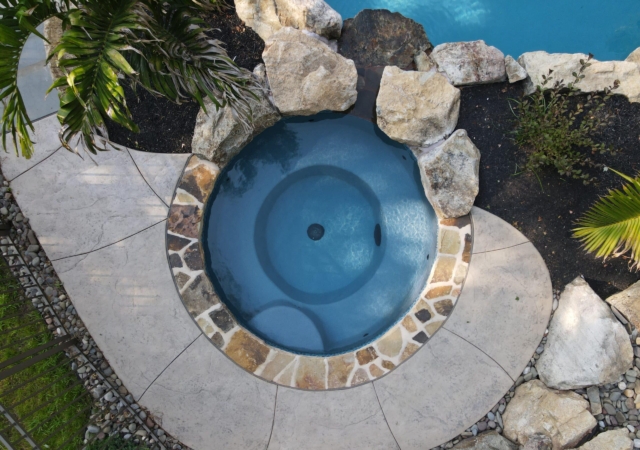 An overhead view of a small, round hot tub with a dark blue bottom, surrounded by large natural rocks and a stone walkway. The hot tub is built into the ground next to a swimming pool, partially visible at the top of the frame.