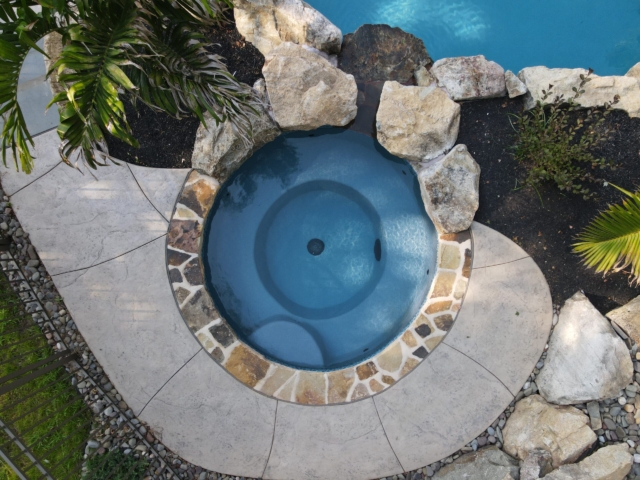An overhead view of a small, round hot tub with a dark blue bottom, surrounded by large natural rocks and a stone walkway. The hot tub is built into the ground next to a swimming pool, partially visible at the top of the frame.