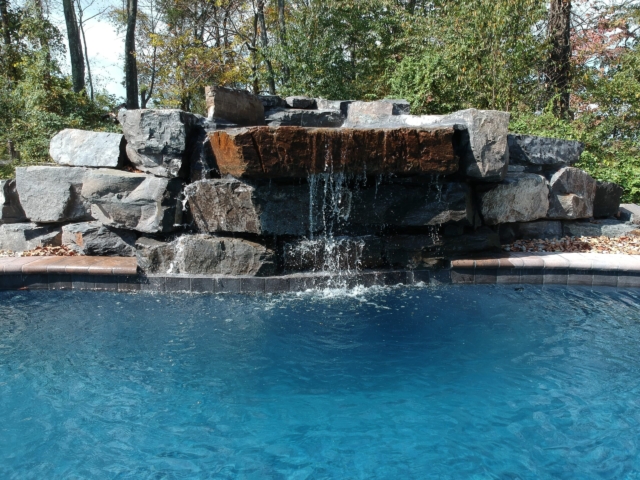 A ground-level shot of a swimming pool with a large, elaborate waterfall feature. The waterfall is constructed from various sizes and colors of natural rocks, with water cascading down into the pool. The pool has a dark blue liner, and its edge is lined with a dark blue tile border and a light brown coping.