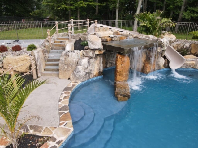 A beautifully landscaped backyard pool area with a natural, freeform pool and a small, built-in slide. The slide is integrated into a rock formation that also features a waterfall flowing into the pool. A set of gray stone steps with a rope handrail leads up to a landing behind the slide. The pool is surrounded by a textured concrete deck with natural stone accents and a blue tiled edge.