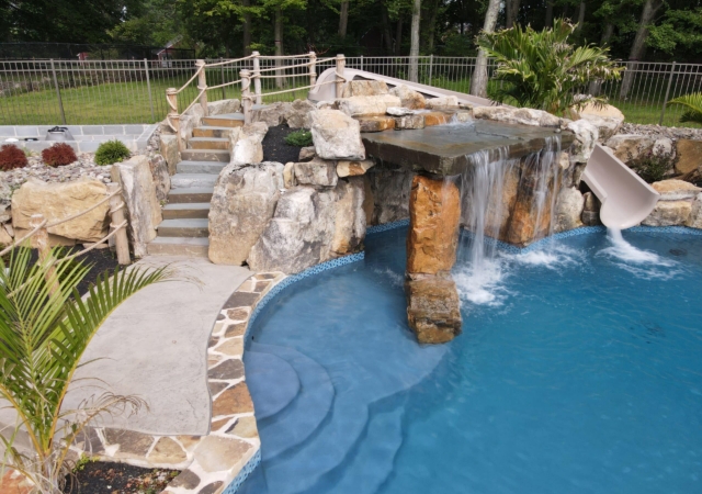 A beautifully landscaped backyard pool area with a natural, freeform pool and a small, built-in slide. The slide is integrated into a rock formation that also features a waterfall flowing into the pool. A set of gray stone steps with a rope handrail leads up to a landing behind the slide. The pool is surrounded by a textured concrete deck with natural stone accents and a blue tiled edge.