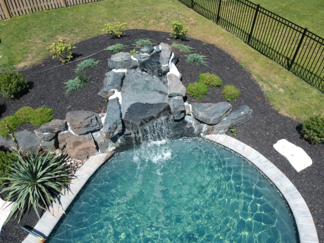 An aerial view of the rounded end of a swimming pool with a small waterfall feature. The waterfall is made of several dark grey rocks and is set into a raised garden bed. The pool has a light blue interior, and its edge is lined with light-colored coping.