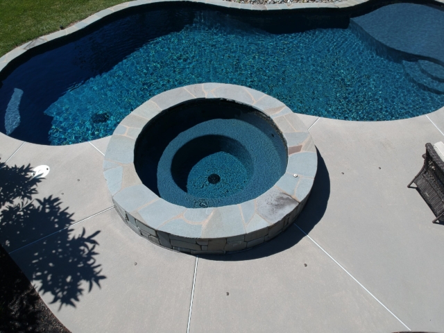 An elevated view of a backyard swimming pool and a circular hot tub. The pool and hot tub both have a dark blue interior and are surrounded by a light grey concrete deck. The hot tub is a raised structure with a stone tile exterior.