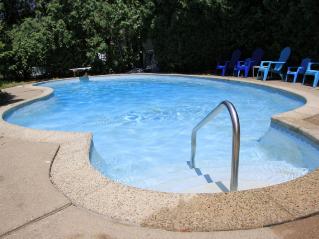 A small, round in-ground swimming pool with a concrete deck is shown. The pool features a set of white steps with a stainless steel handrail for entry. A diving board is visible in the background.