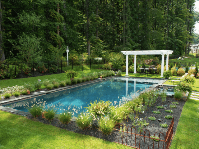 A shot of a rectangular swimming pool with a dark blue interior. The pool is surrounded by a narrow patio and a lush green lawn. A garden area with various plants and a low metal fence is in the foreground.