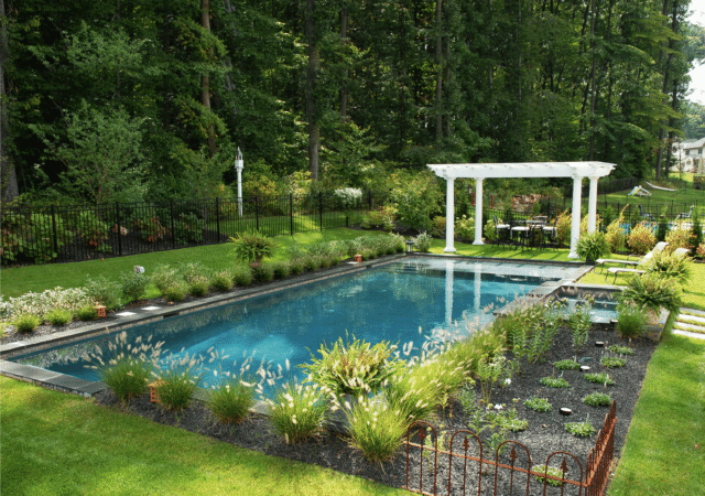 A shot of a rectangular swimming pool with a dark blue interior. The pool is surrounded by a narrow patio and a lush green lawn. A garden area with various plants and a low metal fence is in the foreground.