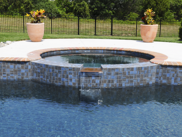 A raised, circular hot tub with a small waterfall feature that spills into a swimming pool. The hot tub's exterior is covered in small, blue and black square tiles. Its coping is a rustic, red brick material.