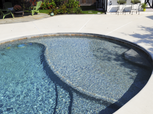 A view of a kidney-shaped swimming pool with a light blue interior. The pool has a curved entry area with a shallow sun shelf. The pool's interior edge is lined with small, multi-colored tiles. The surrounding deck is a clean, white concrete.