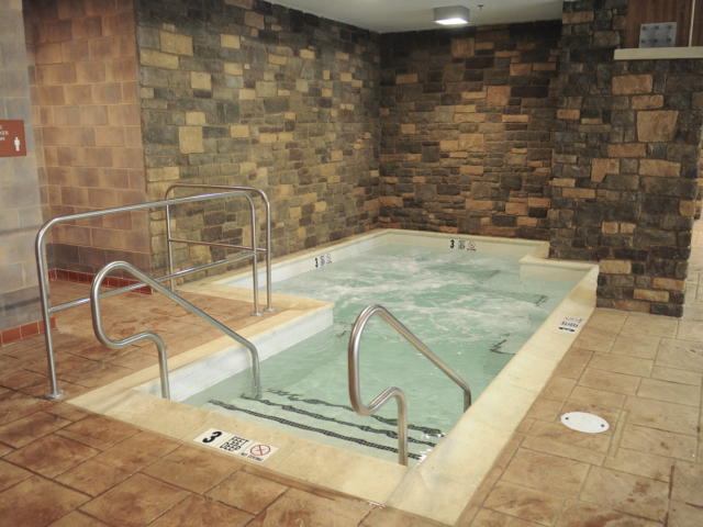 An indoor hot tub with bubbling water and a light-colored tiled edge. The hot tub has two stainless steel handrails for entry and is surrounded by a brown tiled floor. The walls are a mix of different types of stone-like tile.