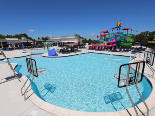 A community water park with a large, freeform swimming pool featuring three basketball hoops along the water. In the background, there is a colorful multi-level play structure with several water slides, buckets, and other features. The pool deck is white and is furnished with rows of dark lounge chairs.