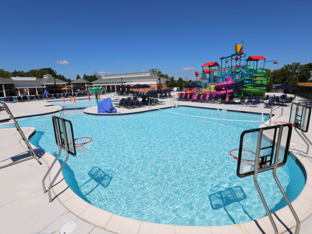 A community water park with a large, freeform swimming pool featuring three basketball hoops along the water. In the background, there is a colorful multi-level play structure with several water slides, buckets, and other features. The pool deck is white and is furnished with rows of dark lounge chairs.
