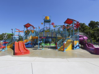 A bright and colorful outdoor water play structure. The structure features multiple levels of platforms, stairs, and railings, all painted in vibrant shades of blue, yellow, and red. There are several slides, including a red one on the left and a purple one on the right, as well as a large yellow bucket at the top that tips over and douses the area with water. The ground is a light-colored concrete splash pad.
