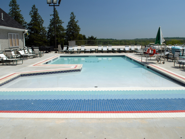 A community swimming pool complex with a large, shallow leisure pool. The leisure pool has a unique tiled bottom with wide stripes of red, white, and various shades of blue. The deck is a light gray concrete and is filled with rows of white lounge chairs and tables.
