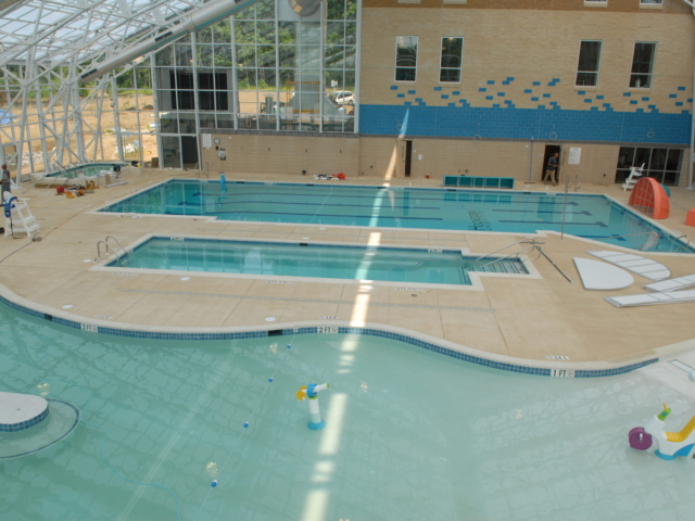 An elevated view of a large, partially constructed indoor aquatic center. There are three swimming pools visible: a large, shallow, freeform leisure pool in the foreground, a rectangular pool with lap lanes in the middle, and a small, square wading pool. The water in the pools is a light blue color. The pool decks are a light brown concrete.