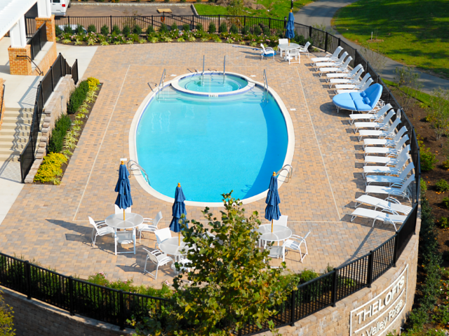 An elevated view of a large, oval-shaped communal swimming pool with a circular hot tub inside one end. The pool deck is made of brick pavers and is furnished with multiple white tables with blue umbrellas, and a long row of white lounge chairs.