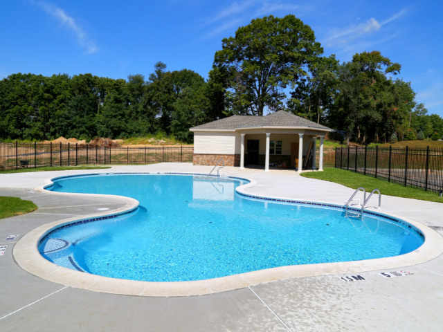 A ground-level shot of a large, free-form community swimming pool. The pool has a light blue interior with a mosaic tile border. The pool deck is a light grey concrete, and two sets of metal handrails provide access to the water.
