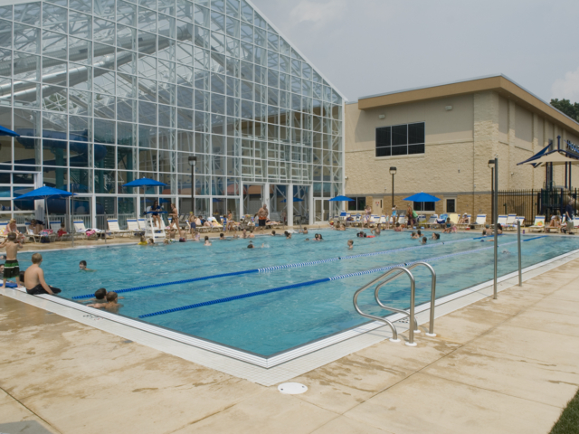 A large, rectangular swimming pool with a bright blue tiled edge and two lap lanes is full of people swimming and enjoying the water. The pool is located outdoors, next to a huge glass-enclosed building, which appears to house an indoor aquatic center with water slides visible through the glass.