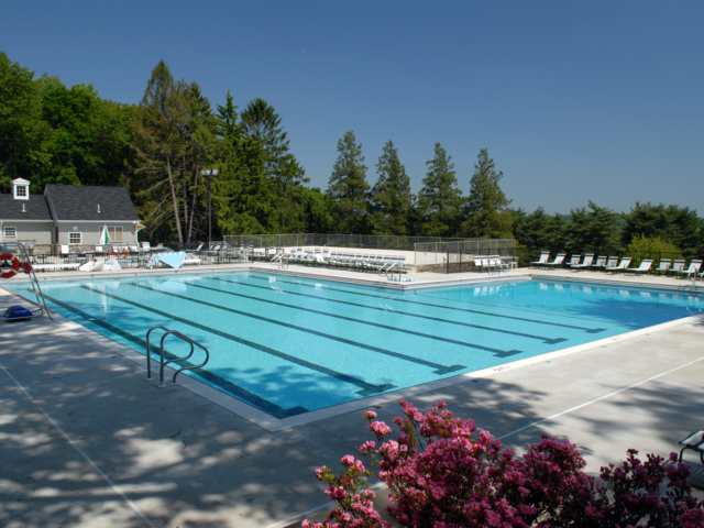 A large, rectangular outdoor swimming pool with five lap lanes is shown on a sunny day. The pool is surrounded by a light-colored concrete deck. The deck is furnished with a large number of lounge chairs.