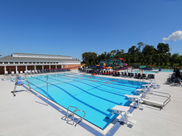 An expansive outdoor aquatic center with a bright, sunny sky. The main feature is a huge, rectangular swimming pool with multiple lap lanes, starting blocks, and diving boards. In the background, there is a colorful water play structure with slides and fountains. The pool deck is white and filled with rows of lounge chairs.