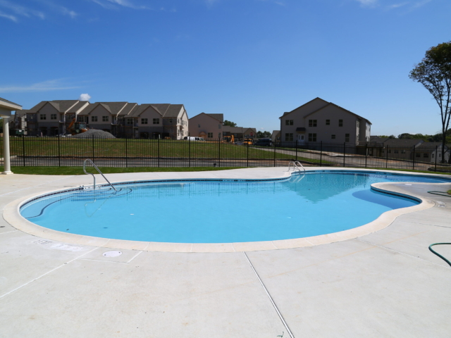 A freeform, kidney-shaped swimming pool with bright blue water is surrounded by a plain concrete deck. The pool is located within a residential development, with several two-story houses and construction sites visible in the background.
