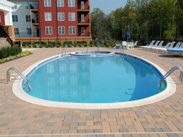 A communal swimming pool complex with a large, oval-shaped pool and a small, circular hot tub. The pools are surrounded by a deck made of multicolored brick pavers. The area is furnished with white lounge chairs and tables with a blue umbrella.