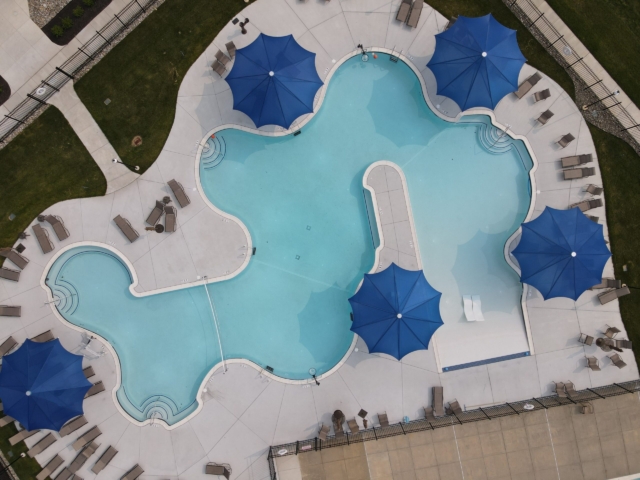 An aerial shot of a large, custom-shaped swimming pool. The main pool has a free-form shape with several connected sections and a light blue interior. It is surrounded by a light-colored concrete deck.