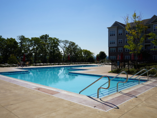 A large, rectangular community swimming pool with a shallow area on one side and a deeper section on the other. A stainless steel handrail leads into the shallow end. The pool is surrounded by a wide concrete deck.