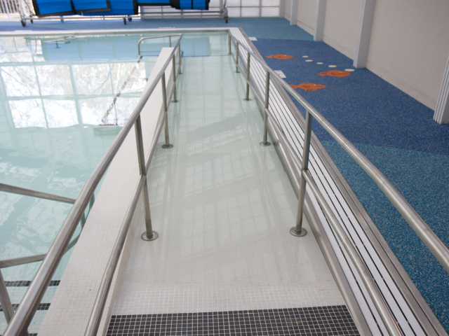 An accessibility ramp with handrails leading into an indoor swimming pool. The ramp is made of a light-colored, non-slip material and is surrounded by a checkered tile pattern at the entrance.