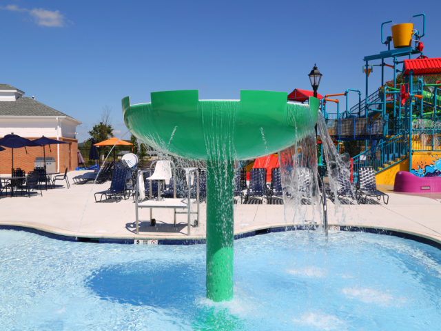 A green mushroom-shaped water fountain with a large bucket spilling water into a swimming pool. The water spills from the mushroom's cap, creating a gentle cascade. In the background, there is a vibrant and colorful water playground structure with slides and a yellow tipping bucket.