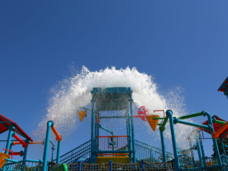 A large bucket on a water park structure tilts, spilling a massive amount of water that splashes out in a wide arc. The structure is made of colorful red, blue, green, and yellow pipes and platforms.