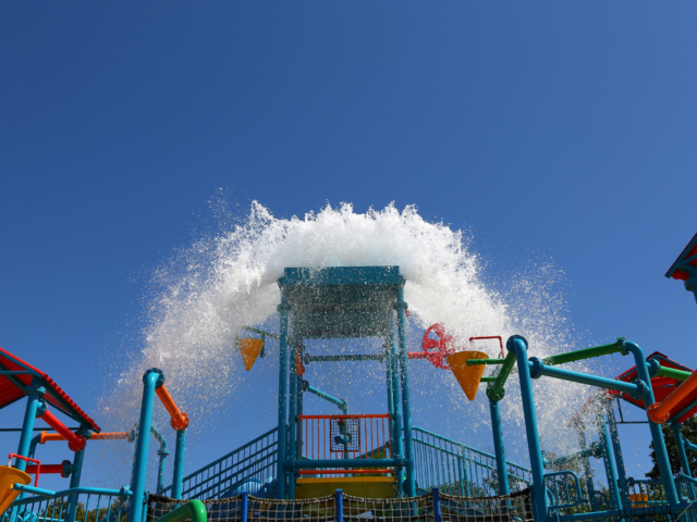 A large bucket on a water park structure tilts, spilling a massive amount of water that splashes out in a wide arc. The structure is made of colorful red, blue, green, and yellow pipes and platforms.