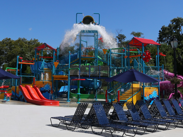 A colorful, multi-level water park playground with slides, ladders, and various water features. A large bucket at the top is tipping over, spilling a large amount of water. The structure features red, blue, green, and yellow elements. In the foreground, there are rows of dark blue lounge chairs and umbrellas on a white deck.