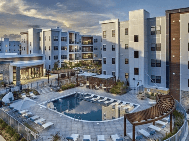 An evening shot of a modern apartment complex courtyard with an inground swimming pool as the centerpiece. The pool is surrounded by a tiled deck with multiple lounge chairs, some under a wooden pergola. A circular metal fence encloses the pool and deck.