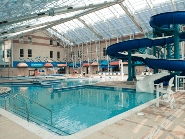 An indoor aquatic center with a large swimming pool and a separate, smaller shallow pool. A large blue and white water slide complex dominates the right side of the image. The building has a high, arched glass and steel roof, allowing natural light to fill the space.