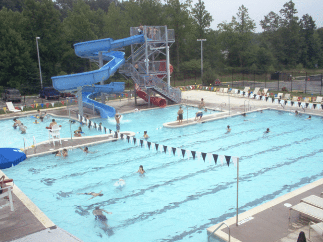 A large, multi-section swimming pool complex on an overcast day. The main pool has lap lanes with several people swimming. In the background, a large water slide structure features two blue spiral slides and one red slide, all leading into a separate splash pool area with several people in it.