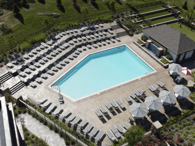 An elevated view of a large, geometric-shaped swimming pool with a light blue interior. The pool is surrounded by a vast concrete deck with a tiered arrangement of gray lounge chairs.