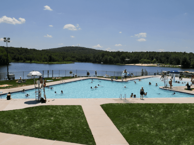 A panoramic shot of a large, arrow-shaped public swimming pool filled with people on a sunny day. The pool is surrounded by a concrete deck with lounge chairs. Beyond the pool area is a large lake with a sandy beach and a forested shoreline.