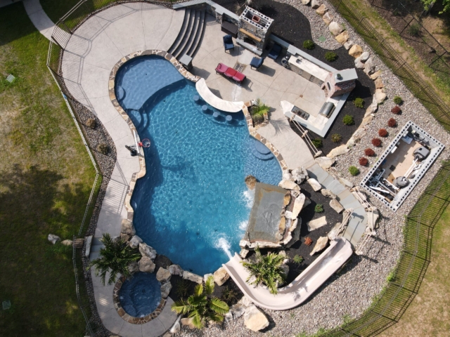 An aerial view of an elaborate backyard swimming pool. The pool has a dark blue liner and an irregular shape. The area is a recreational paradise with features like a waterfall made of large rocks, a water slide, a built-in basketball hoop, and a diving rock. The deck is a light-colored concrete, and there is an outdoor kitchen area, a fire pit, and a separate seating area with red outdoor furniture.