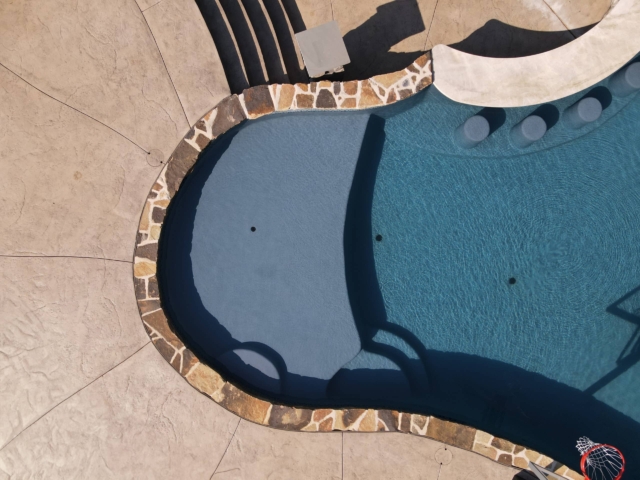 An overhead shot of a swimming pool sun shelf. The pool has a dark blue liner, and its edges are lined with a decorative, multi-toned stone coping. The surrounding deck is a light brown, stamped concrete. In the shallow end of the pool, there are built-in stools, and a small, raised platform with a lighter-colored stone border.