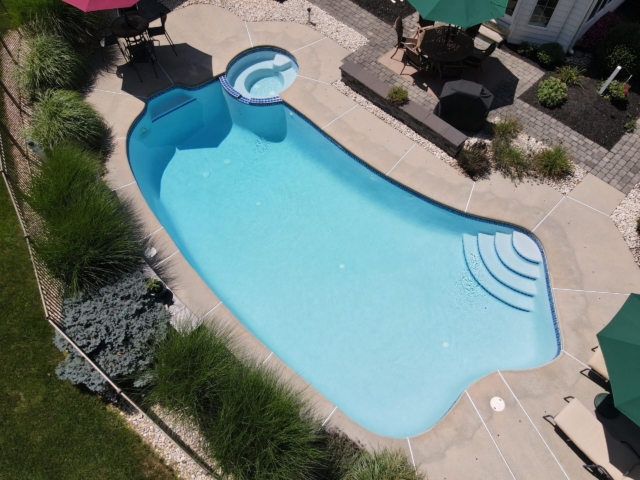 An aerial view of a backyard with a freeform swimming pool and a connected hot tub. The pool's surface is a light blue, and it is surrounded by a light-colored concrete deck. A variety of outdoor furniture and umbrellas are visible on the patio.